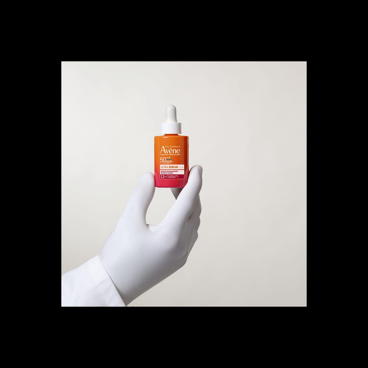 Avène Ultra Serum, gloved hand holding the dropper bottle, with a black border