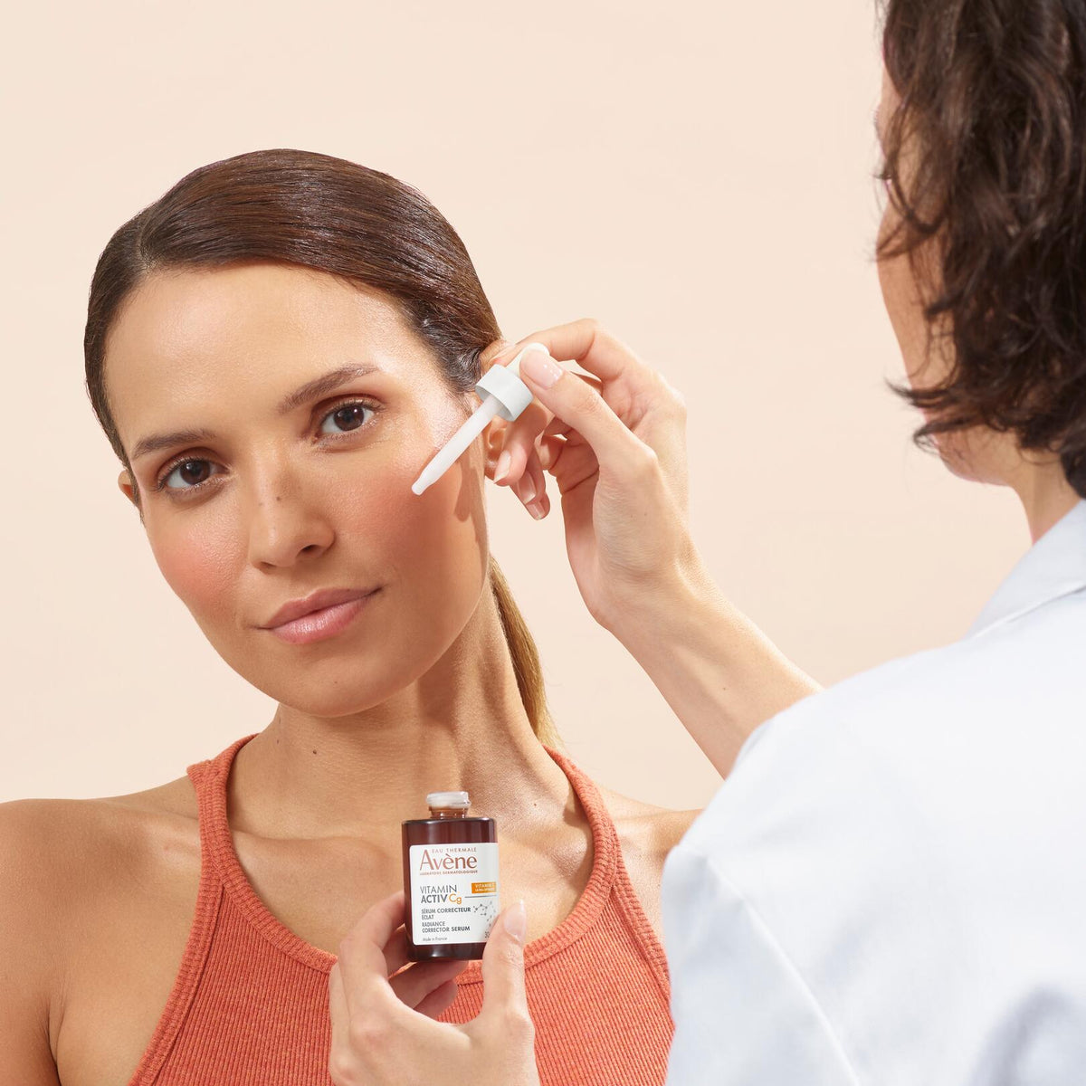 Avène Vitamin Activ Cg, dropper at a woman's cheek next to a person in a lab coat
