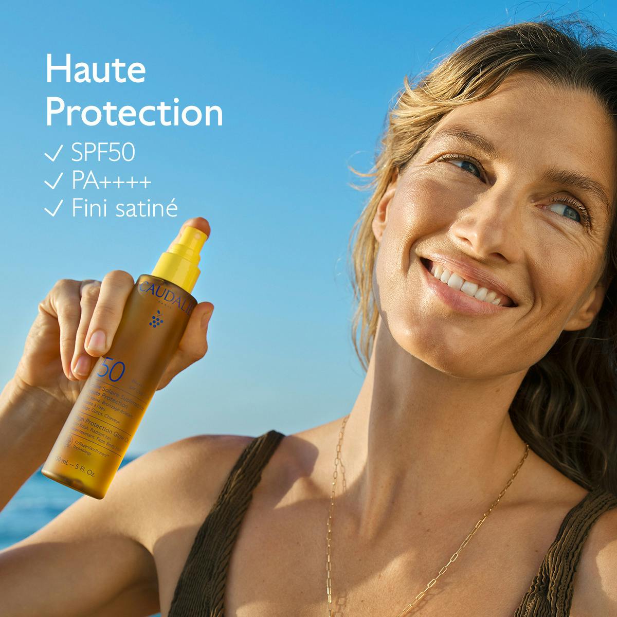 Caudalie Sublimating Sun Oil, smiling woman holding the spray bottle by the sea