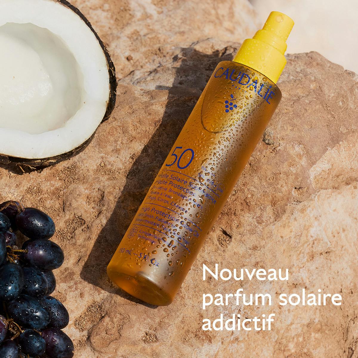 Caudalie Sublimating Sun Oil, spray bottle on a rock with a coconut and black grapes