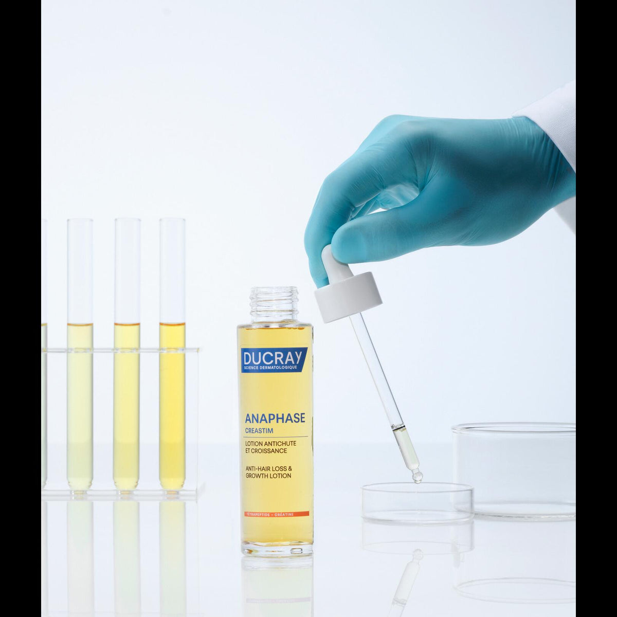 Ducray Creastim, glass bottle next to test tubes and a gloved hand holding the dropper