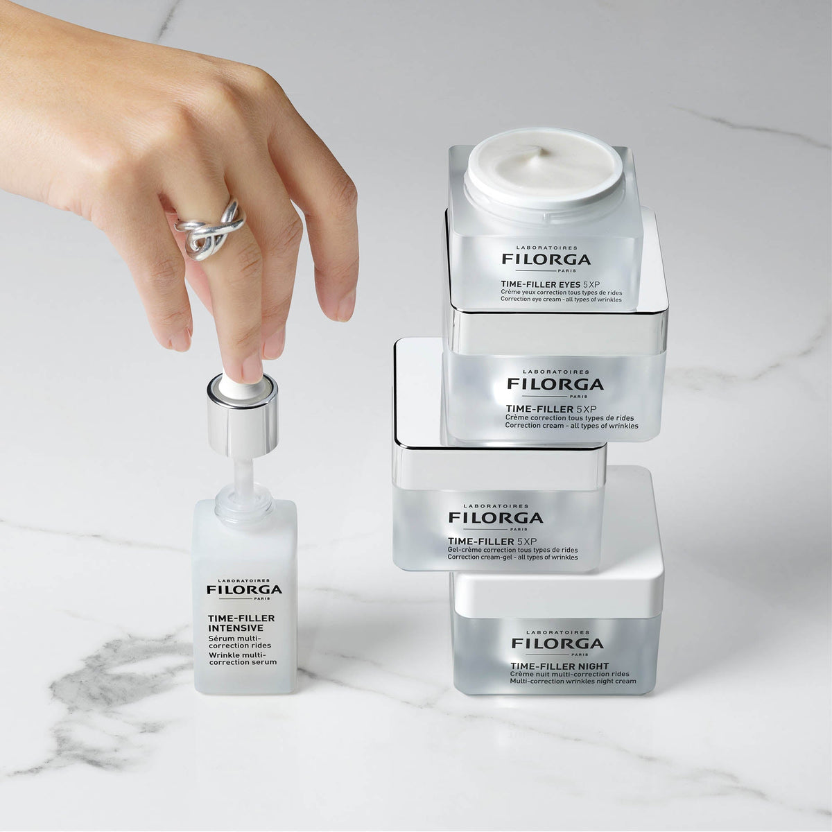 Filorga Time-Filler, hand on a serum bottle beside four stacked jars on marble