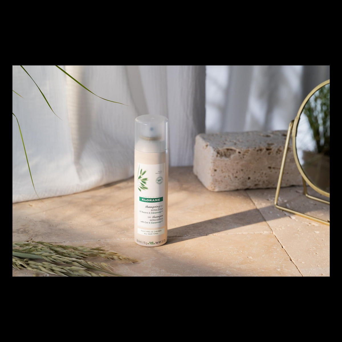 Klorane Oat Milk Dry Shampoo, spray can on travertine tiles beside a round mirror