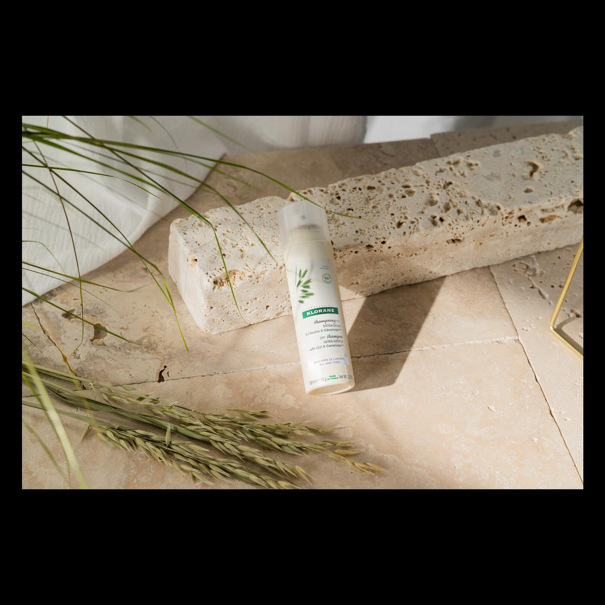 Klorane Oat Milk Dry Shampoo, spray can leaning on a travertine block with oat stems