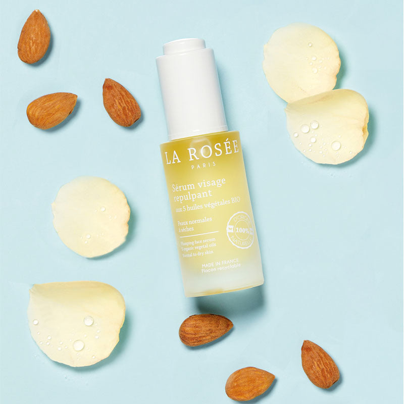La Rosée Plumping Serum, bottle with almonds and petals on a pale blue background