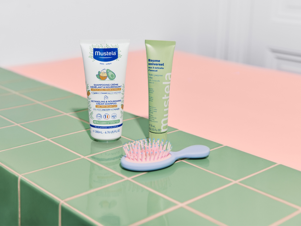 Mustela Detangling Cream Shampoo, tube, green tube and baby hairbrush on green tiles