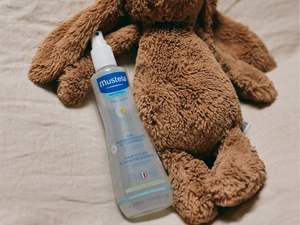 Mustela Refreshing and Styling Water, spray bottle beside a plush teddy bear