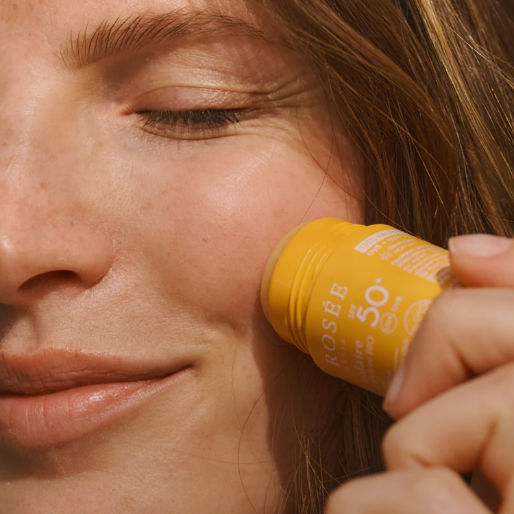 Person applying a yellow sunscreen stick to their face with text about SPF 50+ sun protection.