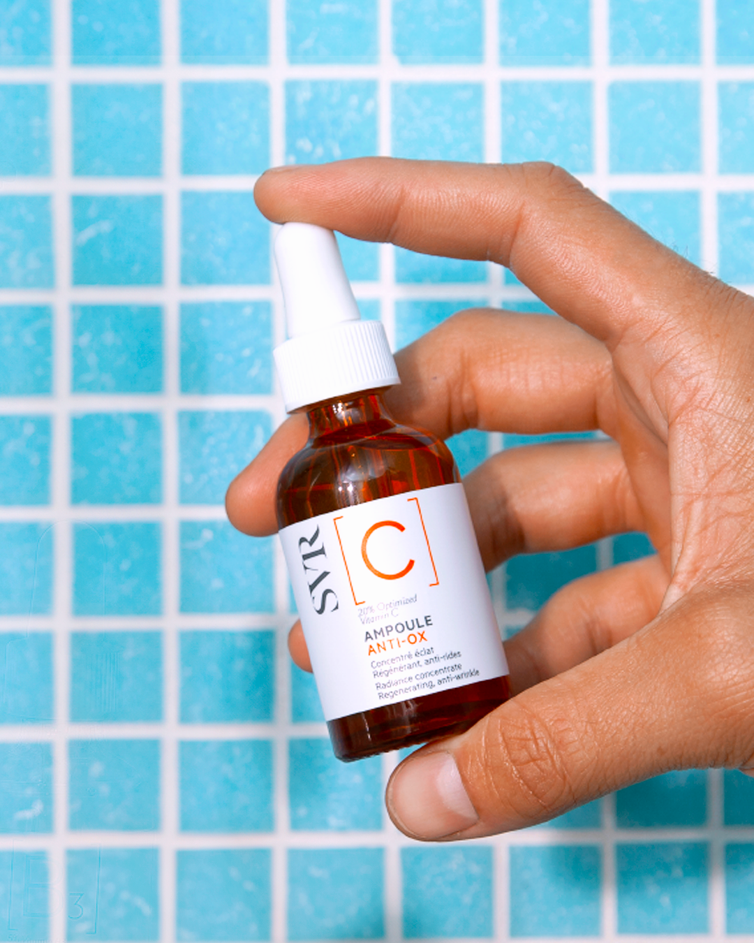 SVR [C] Ampoule Anti-Ox, hand holding the amber glass bottle against blue tiles
